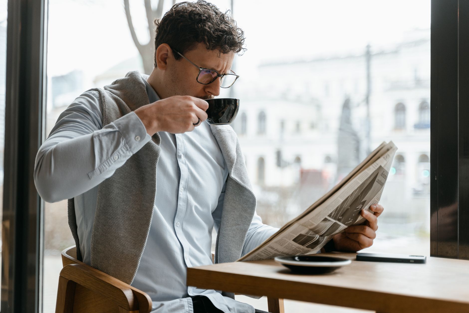 Adult man wearing eyeglasses sipping coffee and reading a newspaper in a cozy cafe setting. — Incognito Casino comment retire